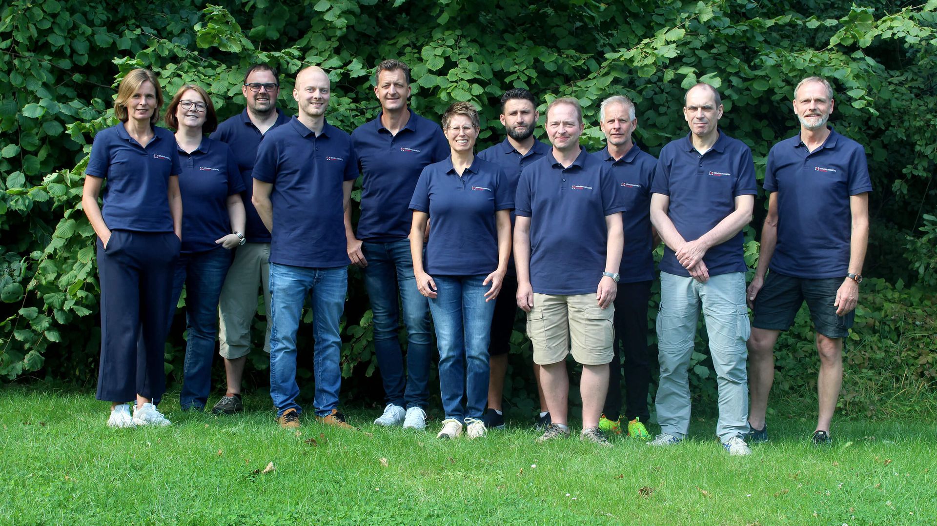 Gruppenfoto des Teams vom Vermessungsbüro Klasen, aufgenommen im Grünen vor einer dichten Hecke. Die zehn Mitarbeiter tragen einheitliche blaue Firmenpoloshirts und stehen entspannt auf einer Wiese, lächelnd in die Kamera.